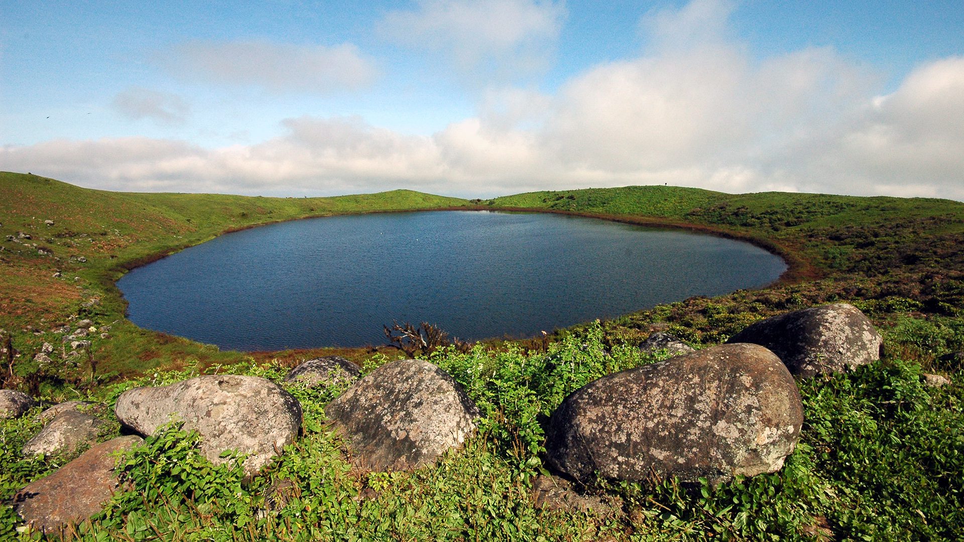 Laguna circular en un cráter volcánico de las islas Galápagos, rodeada de colinas verdes y grandes rocas en primer plano.