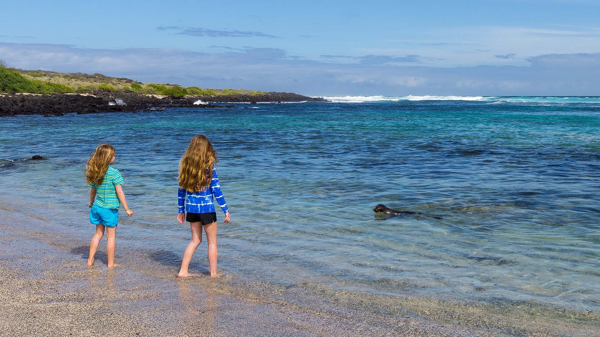 Two children standing on the beach in Galápagos watching a sea lion swim near the shore, representing family-friendly wildlife encounters in Ecuador.